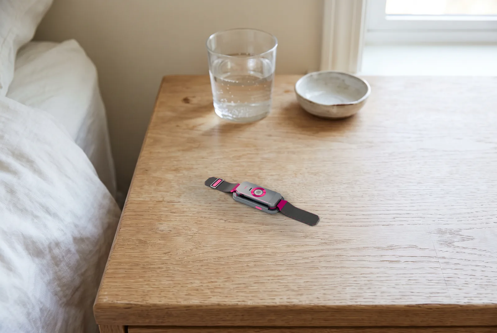 The docked band on a pale oak nightstand in morning light, a glass of water and a small ceramic dish behind it, white linen at the edge of the frame.