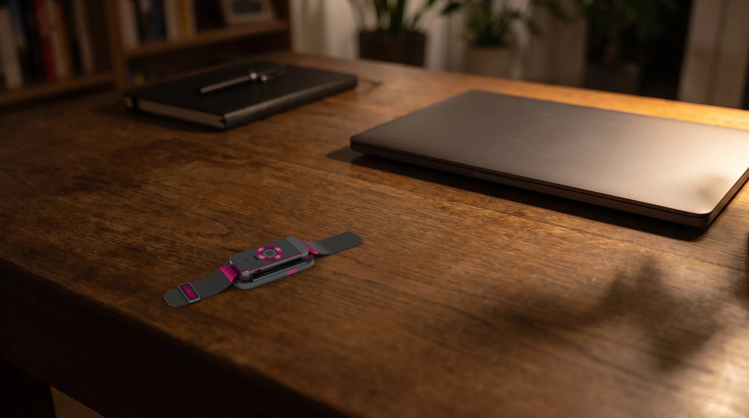 The docked band on the near corner of a dark oak desk in the evening, a closed laptop and a notebook further back under warm lamp light.