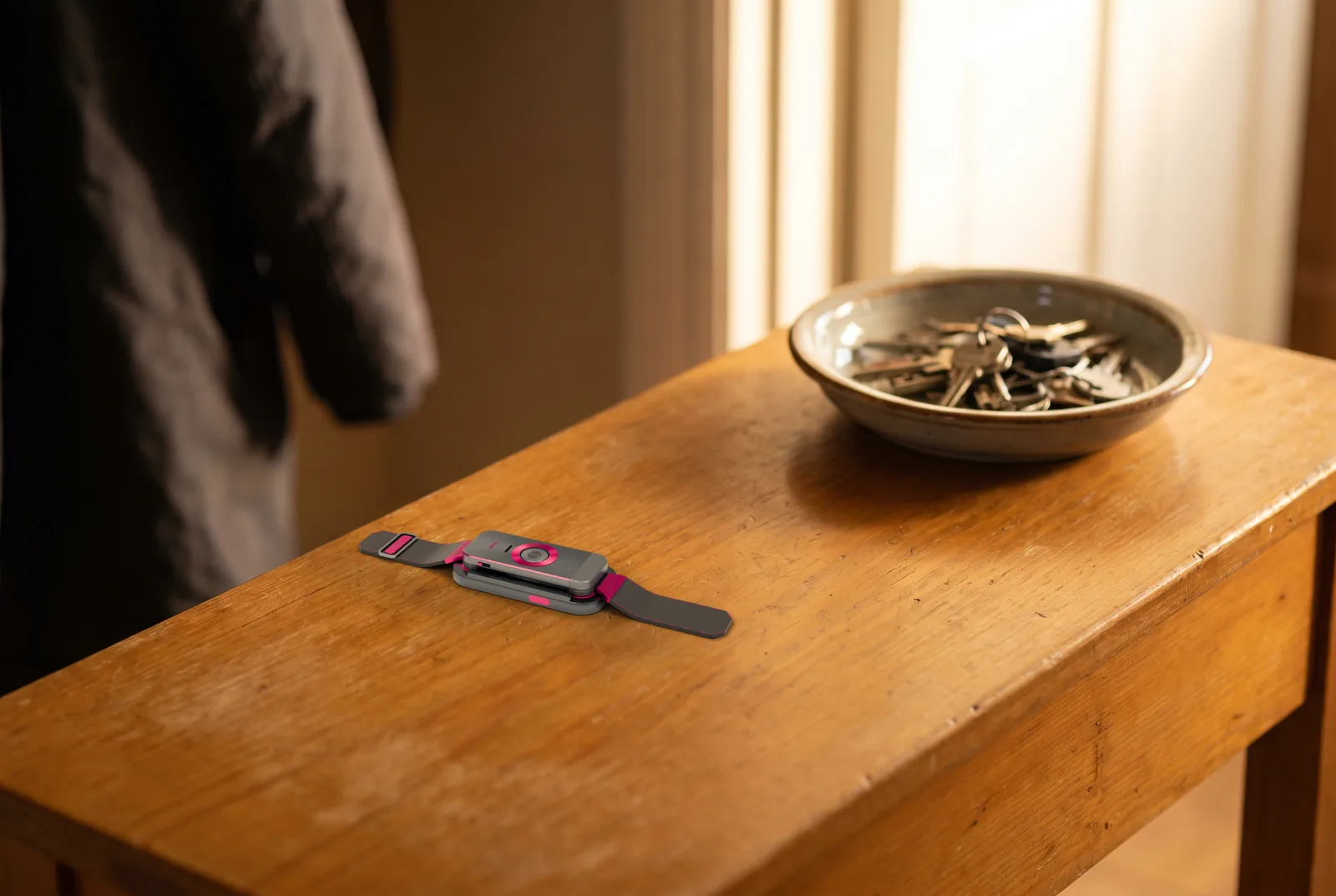 The docked band on a worn honey-coloured hallway console, a ceramic dish of keys behind it and a coat hanging out of focus beyond.