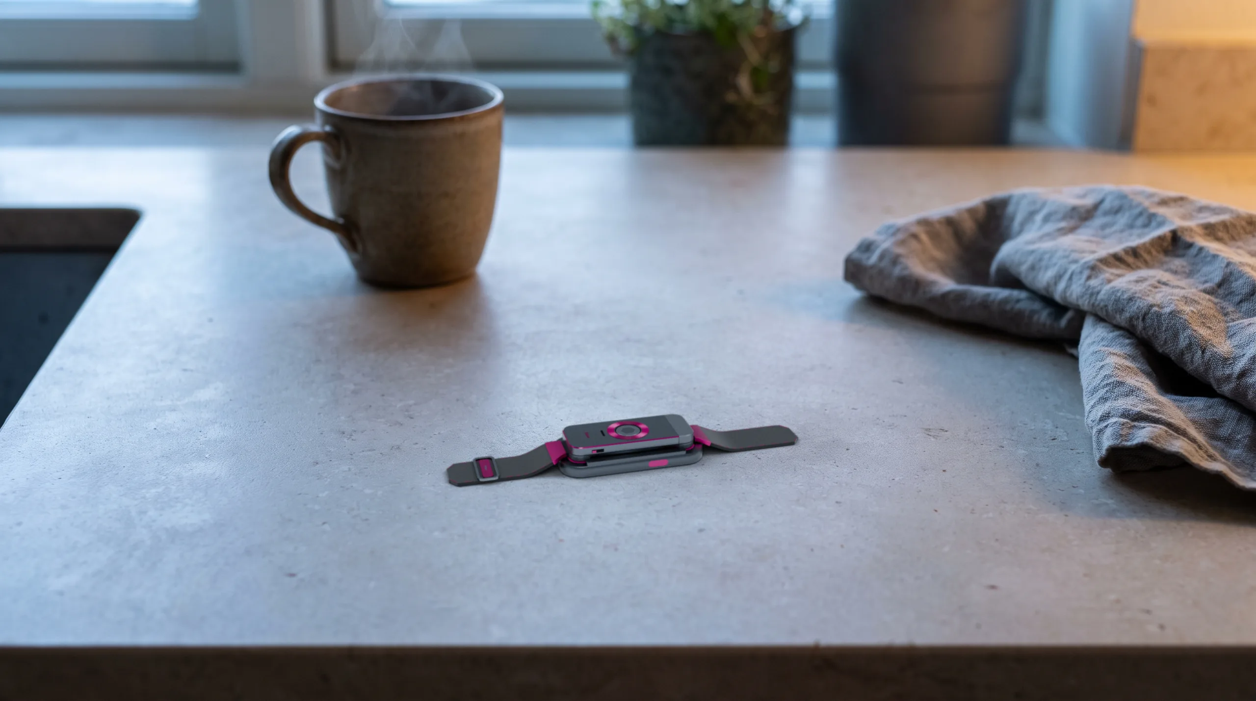 The docked band on a pale stone kitchen counter at dawn, a steaming coffee cup behind it and a folded grey cloth to the right.