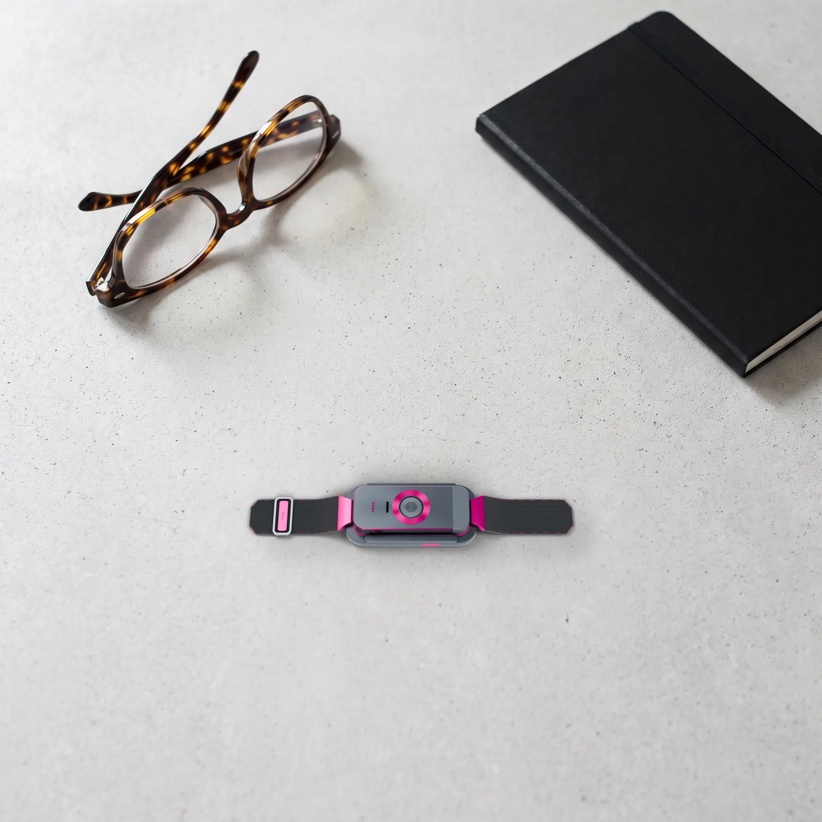 Looking down at the docked band on a pale concrete desk, a folded pair of glasses and a black notebook above it.