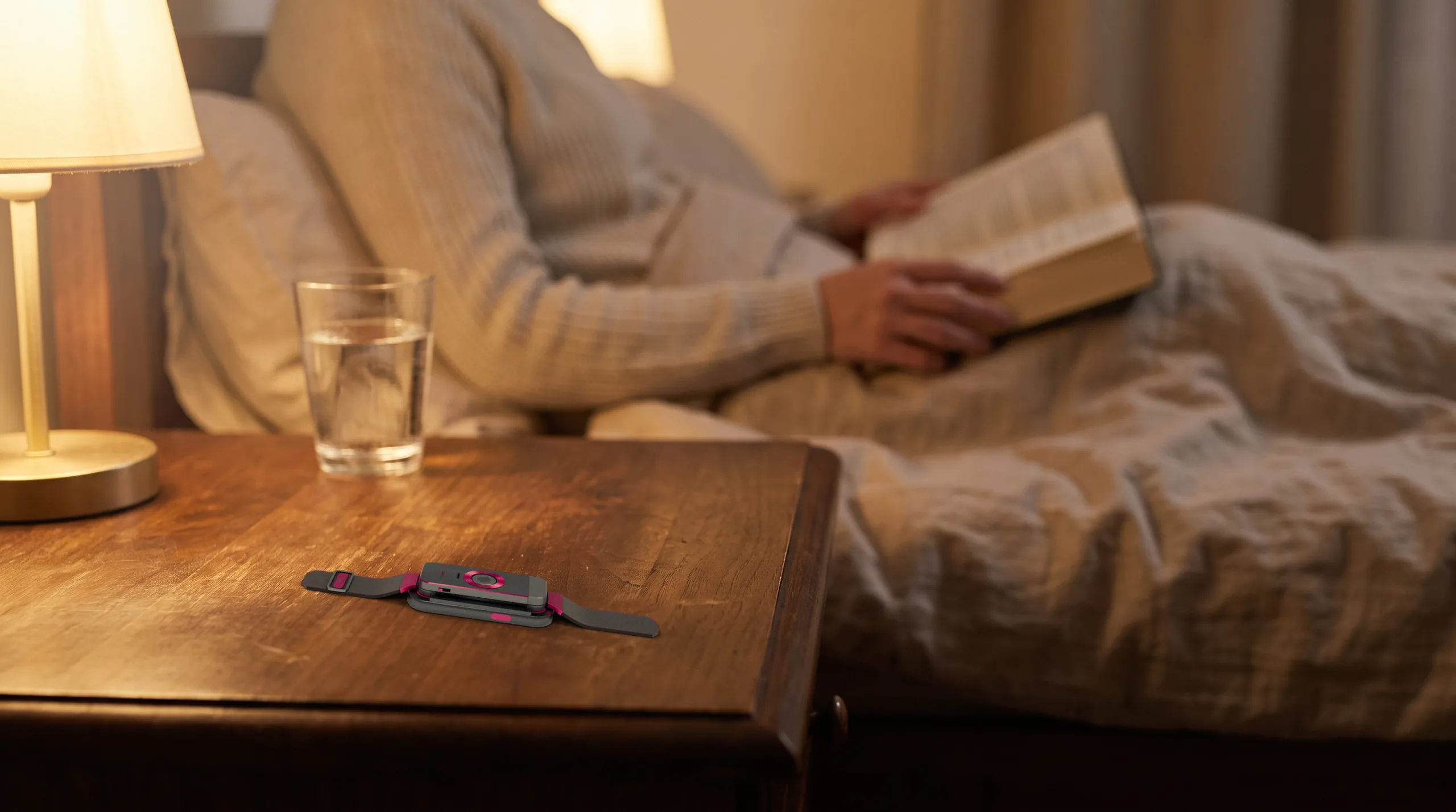 The docked band on a dark walnut nightstand in the foreground; beyond it, out of focus, someone sits up in bed reading under a warm lamp.